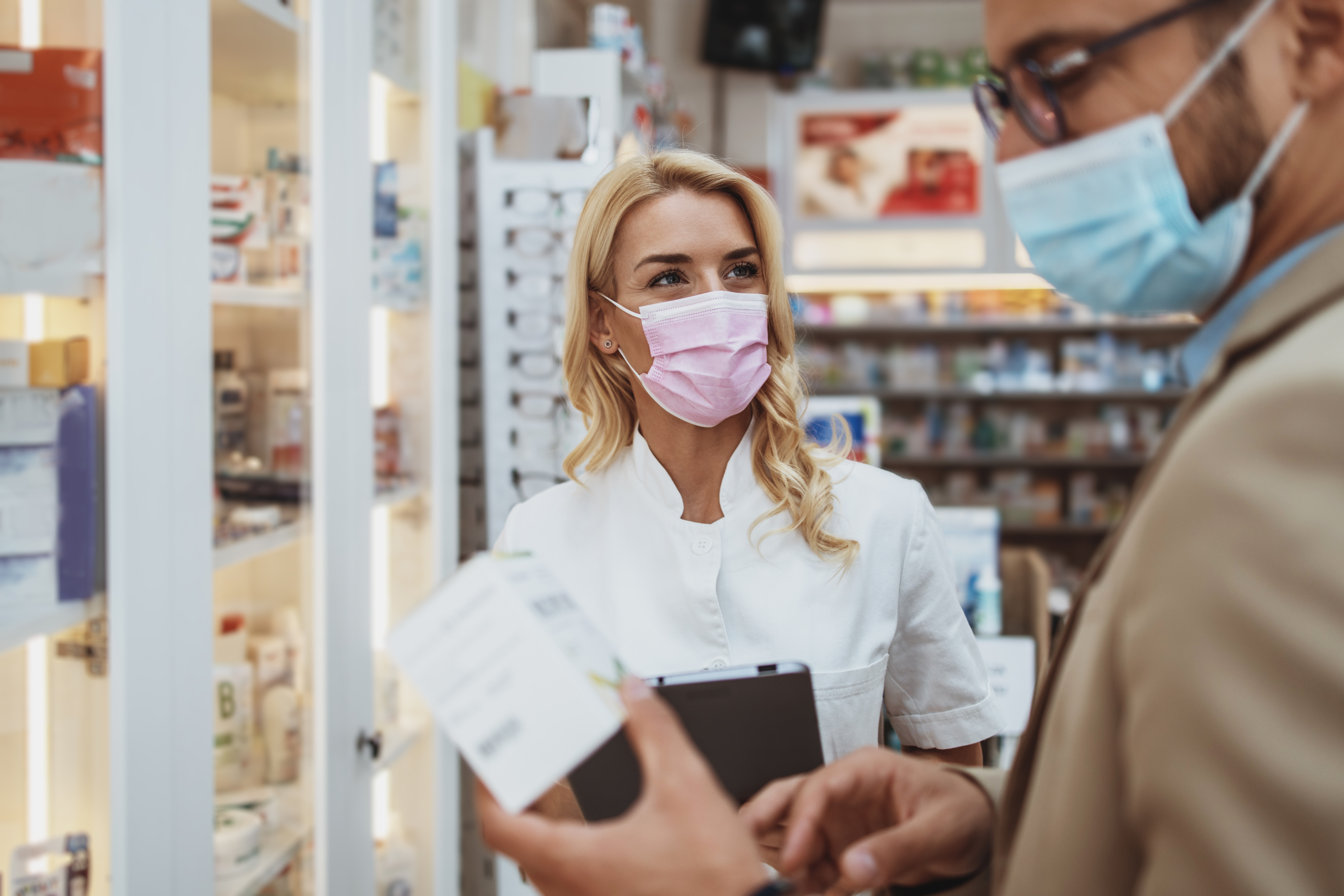Young business man choosing and buying drugs in a drugstore while talking with attractive female pharmacist. She helping him with expert advices. They are wearing protective face masks against virus infection.